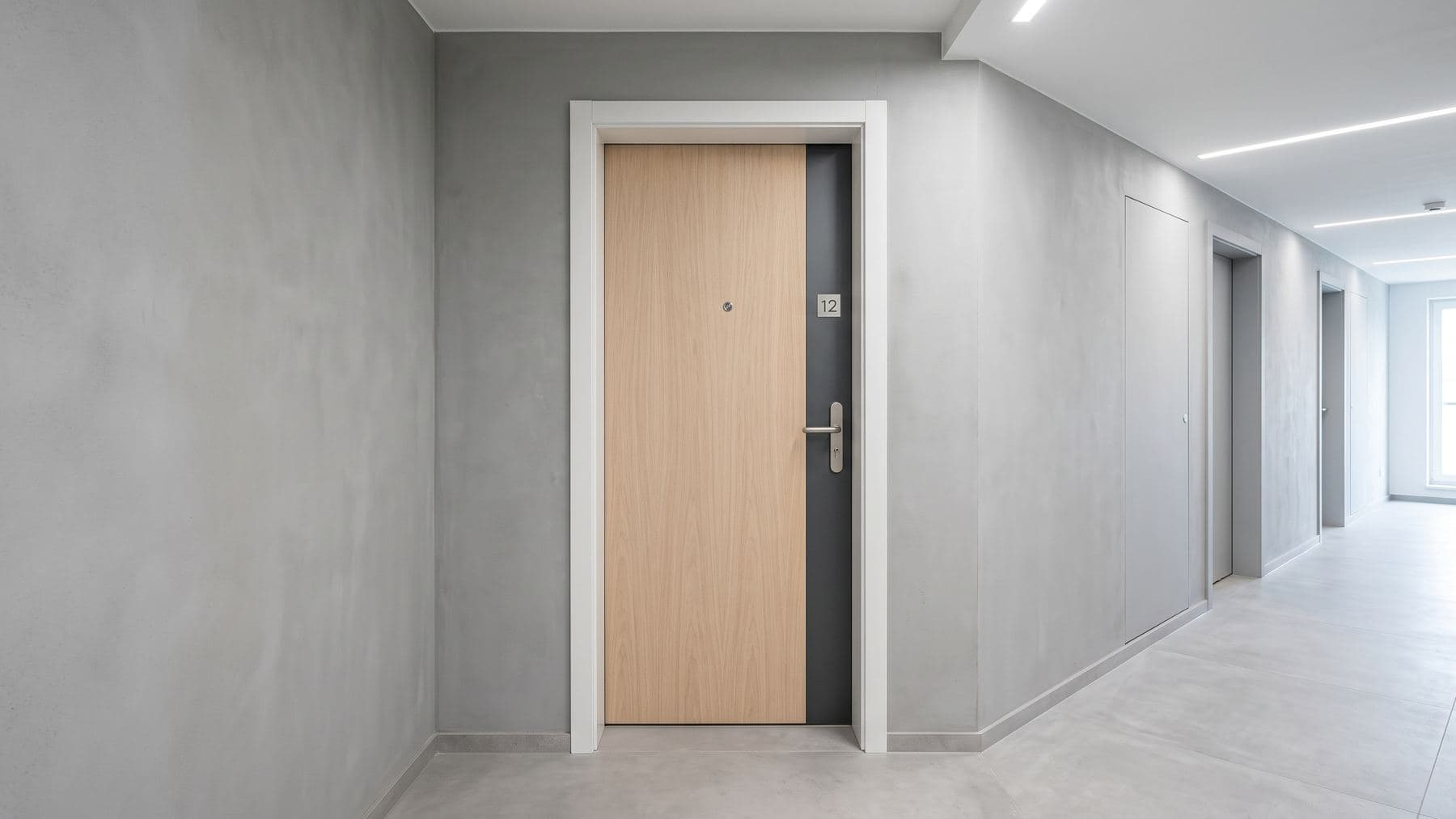 Porte palière d’appartement en chêne clair, plaque anthracite fine et judas, sur un palier au sol lisse