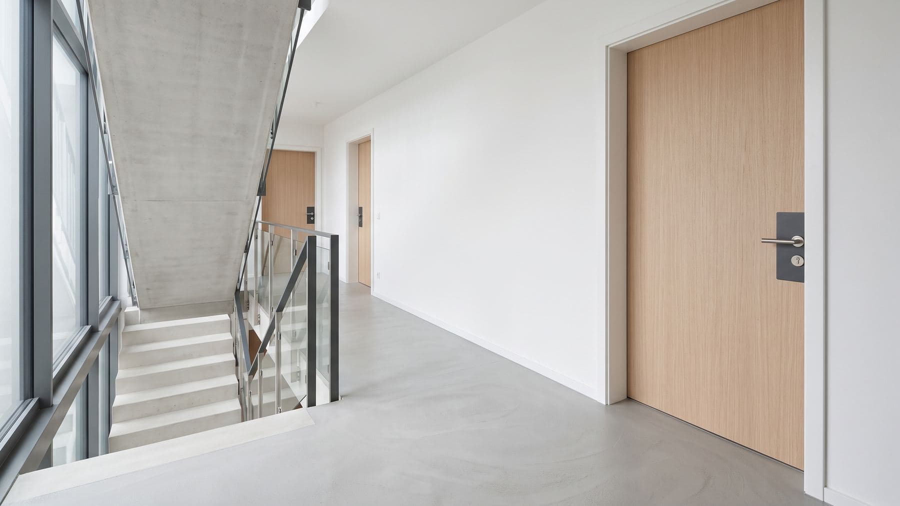 Palier d’immeuble récent vu depuis la cage d’escalier, porte palière en chêne clair et plaque de poignée anthracite